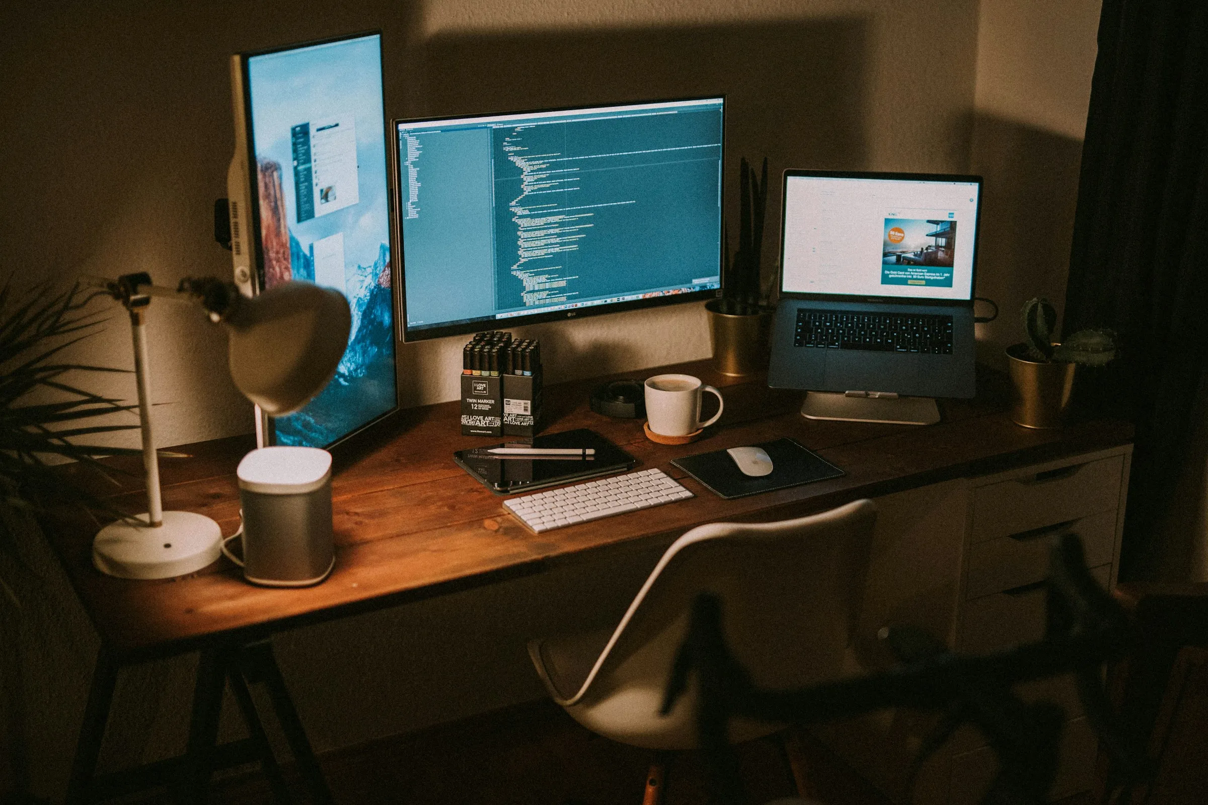 Meja kerja dengan setup monitor berisi kode dan dashboard, diterangi lampu meja